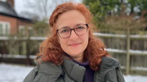 Sam Read/BBC Alex, standing outside, wearing a green coat, with long curly auburn hair. She has glasses on, is smiling and looking at the camera. There is snow behind her, she is standing in front of a fence with a house to the left.