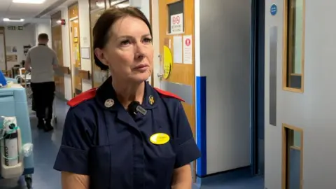 A woman is standing in a hospital corridor wearing a dark blue senior nurse uniform. She has her dark brown hair tied up in a bun and has a yellow ID page on her chest pocket. 