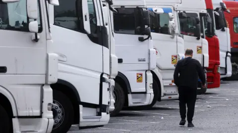 Getty Images Man walking past a line of white lorries with his back to the camera. He is wearing all black.