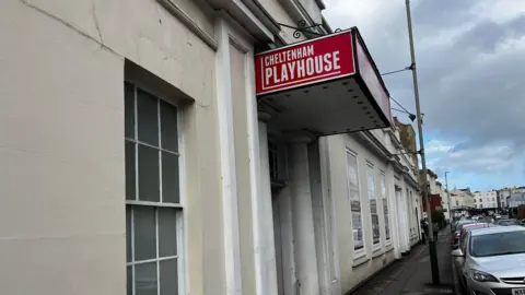 A white Regency style building with a large red sign coming out of it over its door which reads "Cheltenham Playhouse". Cars are parked on the road outside and more buildings can be seen in the distance.