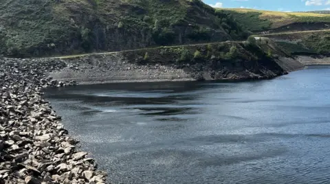 A view from the stone pebble shore at Llyn Brianne. Across the water, the banks of the lake can be seen with horizontal bands of stone emerging out of the water. Beyond the stony banks are hills with grass on them.
