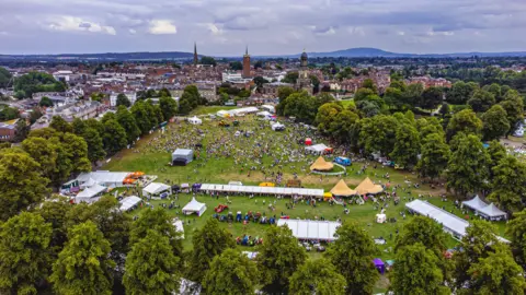 Shropshire Festivals An aerial view of the Quarry Park in Shrewsbury shows trees surrounding a large green field. The field is full of people as well as lines and sections of white stalls, large tents and yellow tee pees. Shrewsbury's skyline s visible in the background