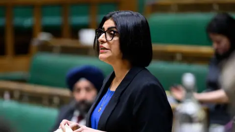 House of Commons Home Secretary Shabana Mahmood in the Commons. She has shoulder-length black hair and is wearing a black suit jacket and blue shirt. A few other MPs are visible in the blurred background.
