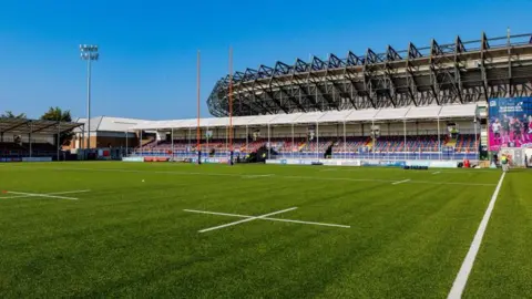 A sunny, empty rugby stadium with a green pitch, goalposts, and seating stands under a bright blue sky.