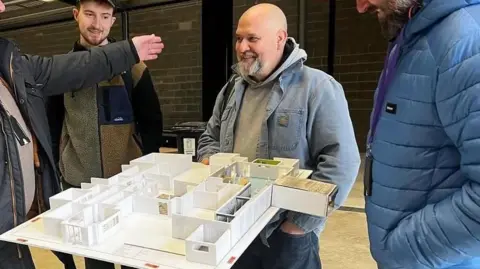 A man smiling with a set in his hands. He has a shaved head and a short, greying goatee beard. He holds in his right hand a board with several small rooms "built" on it out of white material, showing the layout of a building in miniature. He looks to his right towards someone whose arm we can see. 