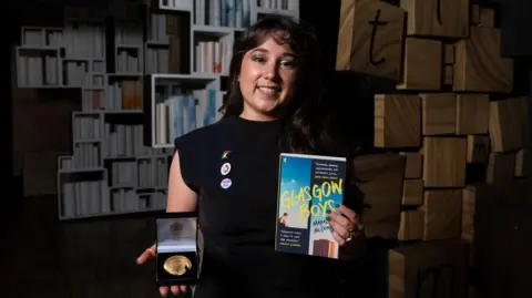 Tom Pilston Margaret McDonald stands in a shadowy room with books on shelves in the background. She holds the Carnegie Medal and her book Glasgow Boys. She smiles at the camera and wears a black top with badges. She has long brown hair and a fringe.