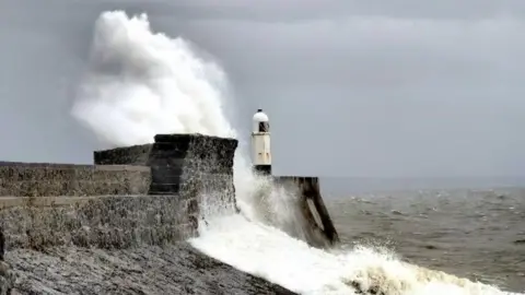 Waves are seen splashing against a harbour wall where there is also a lighthouse 