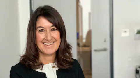 Jersey Police Alison Fossey sits indoors on an office chair,. She has long dark hair and is wearing a black short-sleeved top with white trim and gold buttons. A door and a wall poster are visible in the background.