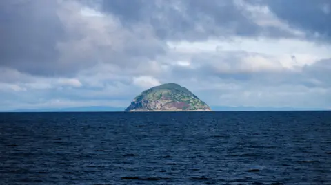 William Dickson A view across dark water towards Ailsa Craig. The water is a dark blue colour. The island is brown and rocky with patches of green. The sky above is blue but streaked with white and grey clouds.