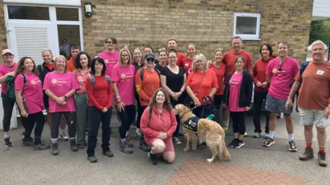Justin Dealey/BBC A group of men and women, wearing pink or red T-Shirts and walking boots or trainers. They are lined up in front of a wall and all of them are smiling. In the front row one of the women is holding the lead of a golden retriever.
