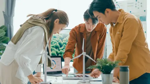 People gathered around a table looking at research.  There is a model windmill on the table. On the left is a woman wearing a white shirt, in the middle a man in an orange jacket and on the right a man in a yellow shirt. 