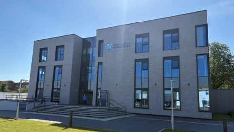 A grey building including the words University of Worcester and its logo. About half a dozen steps lead up to the entrance and grass is on the bottom left of the photo.