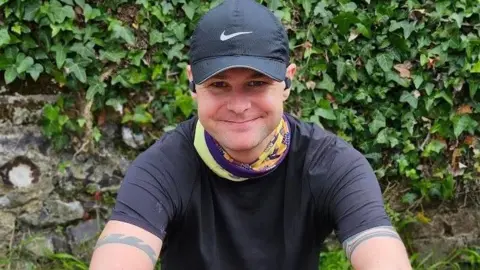 Cameron Taylor smiles at the camera. He is wearing a black tshirt, black hat, earpods and a purple and yellow running scarf.