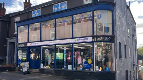 BBC The outside of a blue charity shop called 'St Barnabas'. The shopfront has a navy frame and large glass windows both up and down. Inside there are mannequins clothes and shelves with bric-a-brac in the bottom windows. There is a white sign on the pavement outside. To the right, there is a black sign reading No 25. To the side of the shop are houses, and a large wall painted grey.