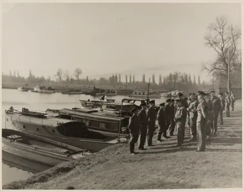 Royal Berkshire Archives A black and white photo shows moored boats to the left and a line of UTP volunteers standing in front of them on the riverbank. Facing them is a line of British Armed Forces officers.