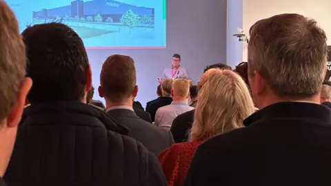 the backs of heads of people listening to a man on a stage speaking