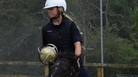 A young woman on a black horse holding a yellow ball wrapped in a metal casing with handles. She is wearing black clothing, a white helmet, and has long blonde hair. She is gazing to the right and in the background are trees and a brown fence.