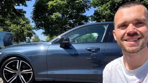A man smiles as he takes a selfie while he is in front of his car - a dark-coloured saloon. 