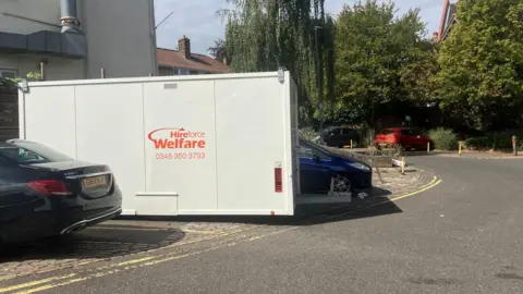 Owen Sennitt/BBC A metal temporary building can be seen in a car park, next to parked vehicles, with the words "Hireforce Welfare" written on it.