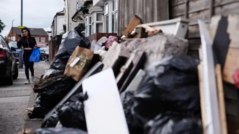 PA Media A big pile of rubbish bags and various other bits of waste piled up on a pavement next to a wooden fence outside of a row of terraced houses. A woman is looking at her phone and holding a blue bag as she walks towards the pile.