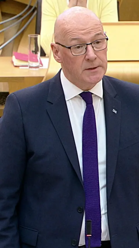 John Swinney wearing dark blue suit and purple tie, speaking in Holyrood chamber after re-appointment