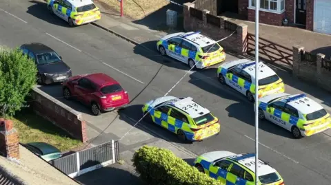 YappApp Overhead view of police cars parked on a residential street, with a tape cordon in place across the road.
