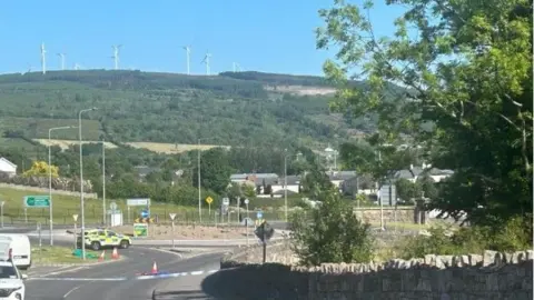 A roundabout, cordoned of with police tape, sits at the foot of a large green hill. On-top of the hill are a handful of wind turbines. A police car sits beside the roundabout, behind the tape. A grey stone wall can be seen running down the road, on the right of the picture, towards to roundabout. 