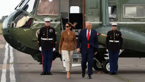 Reuters Two soldiers flank the open doorway of a dark green military helicopter. A woman in cream pants, high heels and a brown suede jacket steps out onto the tarmac, holding the hand of a man in a navy blue suit with a red tie.