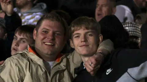 Scott Farrington with his son at a Tamworth FC game. He has his arm around him and is wearing a ring. His son is wearing a Nike jacket