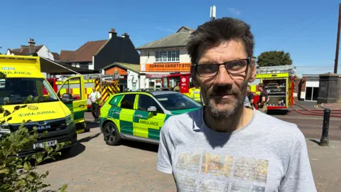 BBC A man with a beard and glasses wearing a grey t-shirt is standing in front of ambulances and fire engines.