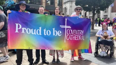 Four people smile as they hold up a rainbow banner which reads Proud to be Coventry Cathedral, with a cross of nails icon in white. To their right a lady in a wheelchair wearing a pink headband smiles broadly, holding a lion teddy. They are in a public square, with others carrying rainbow flags visible behind.