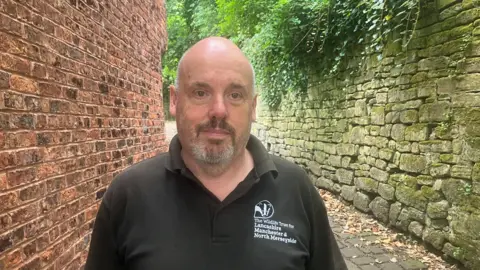 Photograph of Alan Wright from the Lancashire Wildlife Trust. He is pictured on a cobbled footpath next to Rock Hall in the Moses Gate Country Park in Farnworth.