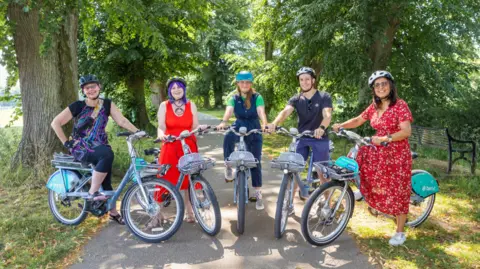 The University of Worcester's Katy Boom, councillors Jill Desayrah and Hannah Cooper, Marcus Chandler from Beryl Bikes and Jas Cartwright of Worcestershire Acute Hospitals NHS Trust celebrating a year of Worcester's Beryl Bike scheme last summer. They are all sat on a bike with helmets on a park path underneath some trees.
