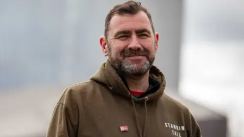 PA Media Man with black and grey beard and dark hair wears a khaki coloured hoody with Standing Tall written on it and a red and white badge which reads the Army Benevolent Fund