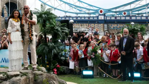 Reuters A woman and a man standing on top of a rock in front of a tree. To the right of the picture is a group of schoolchildren wearing red uniforms. They are standing behind a rope. In the distance is a bridge.