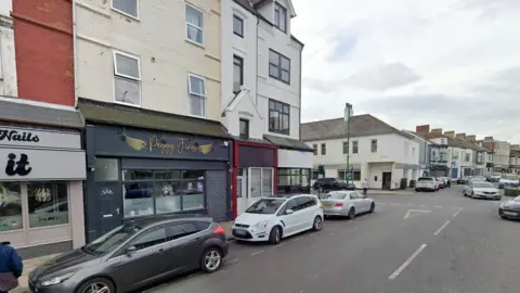 Queen Street has a row of buildings on either side. The bottom floor consists of shop fronts - including the grey shop front for Peggy Junes. The upper floors are residential. A number of cars are parked on the road.