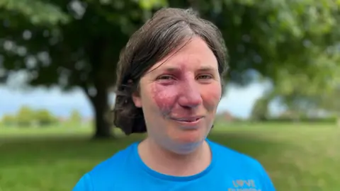 Alice Chambers is wearing a blue T-shirt and has shoulder-length brown hair and is stood in front of trees