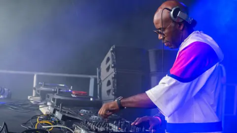 Tracey Welch/BBC DJ Spoony, wearing a baggy T-shirt, glasses and headphones, controls a music mixing desk