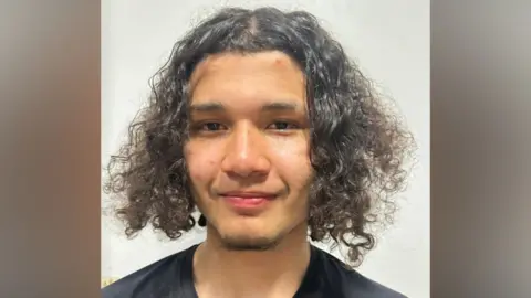 Teenager with long, curly hair smiles at camera - he is wearing a black T-Shirt