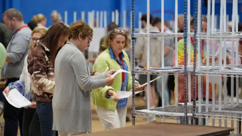 A woman is holding ballots and looking at them. She's next to a shelf full of ballots. She's wearing a long grey cardigan and has red glasses on. A woman next to her has short blond hair with glasses resting on her head. She's wearing a blue top with a yellow cardigan and white trousers. Other people are in the background. 