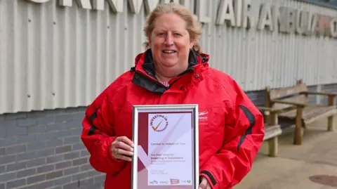 Cornwall Air Ambulance Jackie Eastwood, Cornwall Air Ambulance volunteer manager stood outside looking straight at the camera, wearing a red Air Ambulance coat. She is smiling and holding a framed certificate. 