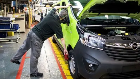 Getty Images A male employee checks the front wheel of a lime green Opel Vivaro medium sized van on the final assembly line at the Vauxhall plant in Luton, UK. He is seen from the back and wears grey cargo trousers and a bottle green T-shirt.