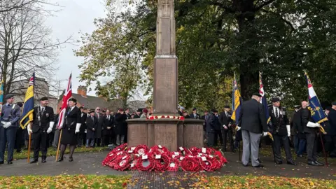People stand with flags at the war memorial which is surrounded by poppy wreaths. Many are wearing uniform.