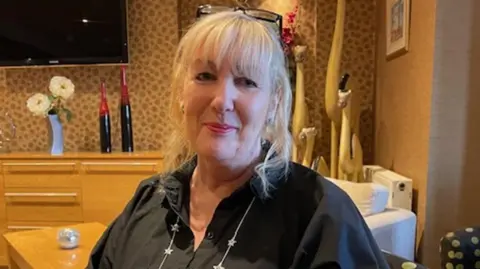 Andy Mitchell/BBC Claire Smith sitting in a hotel room. She is smiling, has long blonde hair and is wearing a black shirt with her glasses on the top of her head