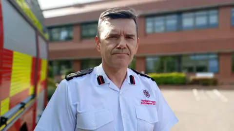 Wright is standing next to a fire engine. he's wearing a white shirt with black lapels and the fire service emblem. A building is in the background. 