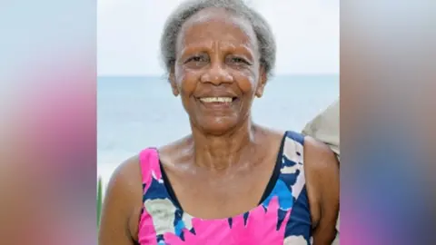 Mel Chevannes photographed by the sea, wearing a pink and blue brightly-coloured sun dress, and looking happy. She has short grey hair and is smiling at the camera.