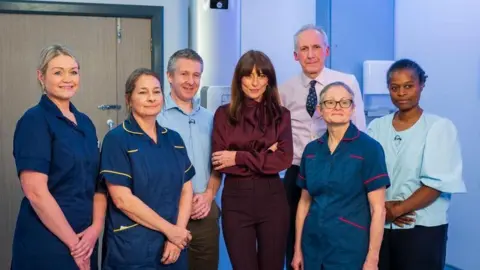 CUH Davina McCall is standing with three women dressed in nurse uniforms, a woman wearing a blue blouse and the men wearing shirts. They are in a hospital.