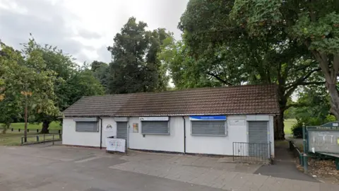 Google A white one-storey building with grey shutters over its windows and doors surrounded by large trees