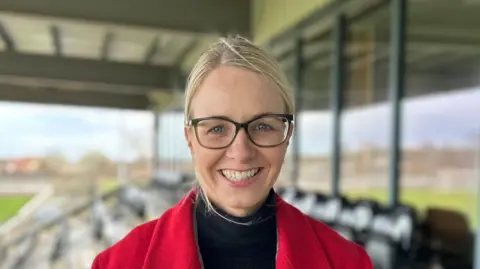 Nicola Sanders is in the stands at Ashington Football club. She has blonde hair tied away from her face and is wearing dark-rimmed glasses and a red coat. 
