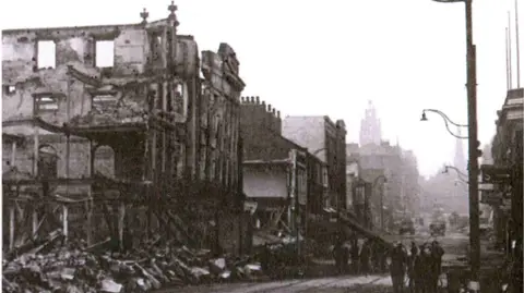 A black and white picture shows a street filled with bombed buildings and rubble. Several people appear to be standing on the right, huddled in a small group.